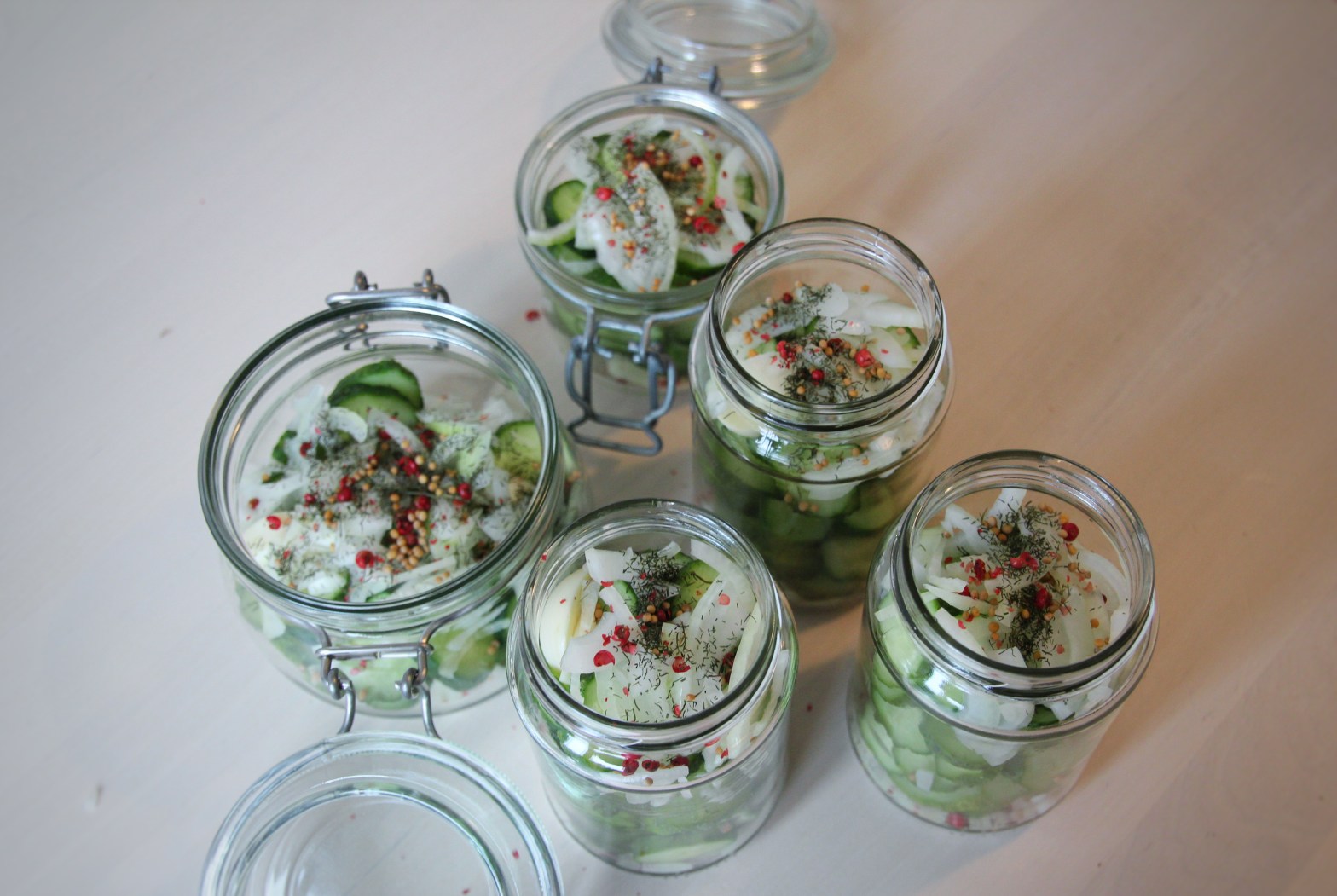 Five open jars of pickles seen from the top, inside are some green cucumbers topped with sliced onion and spices