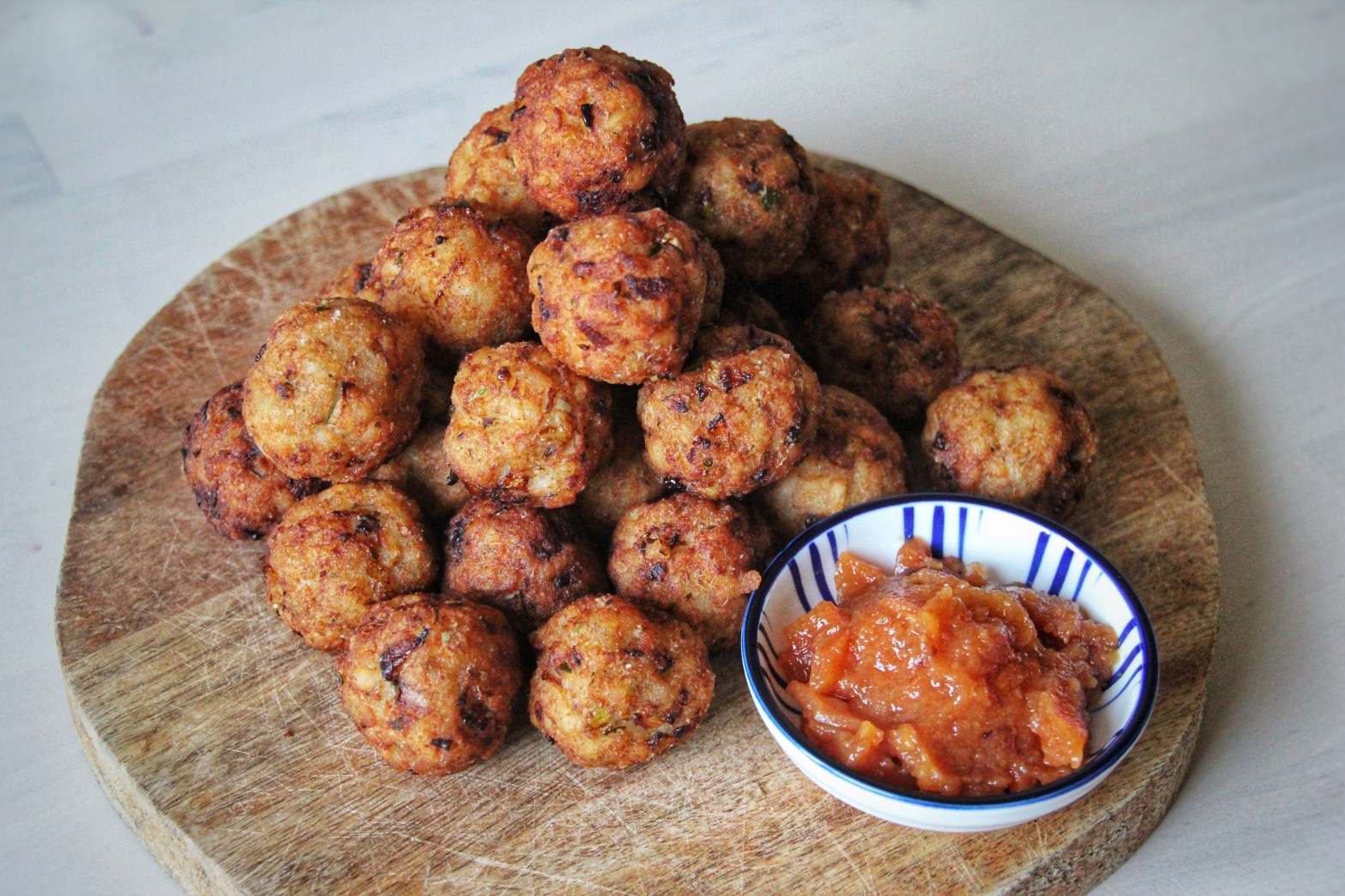 A pyramid of deep fried balls on a wooden plank, with a small bowl of chutney in the bottom right corner