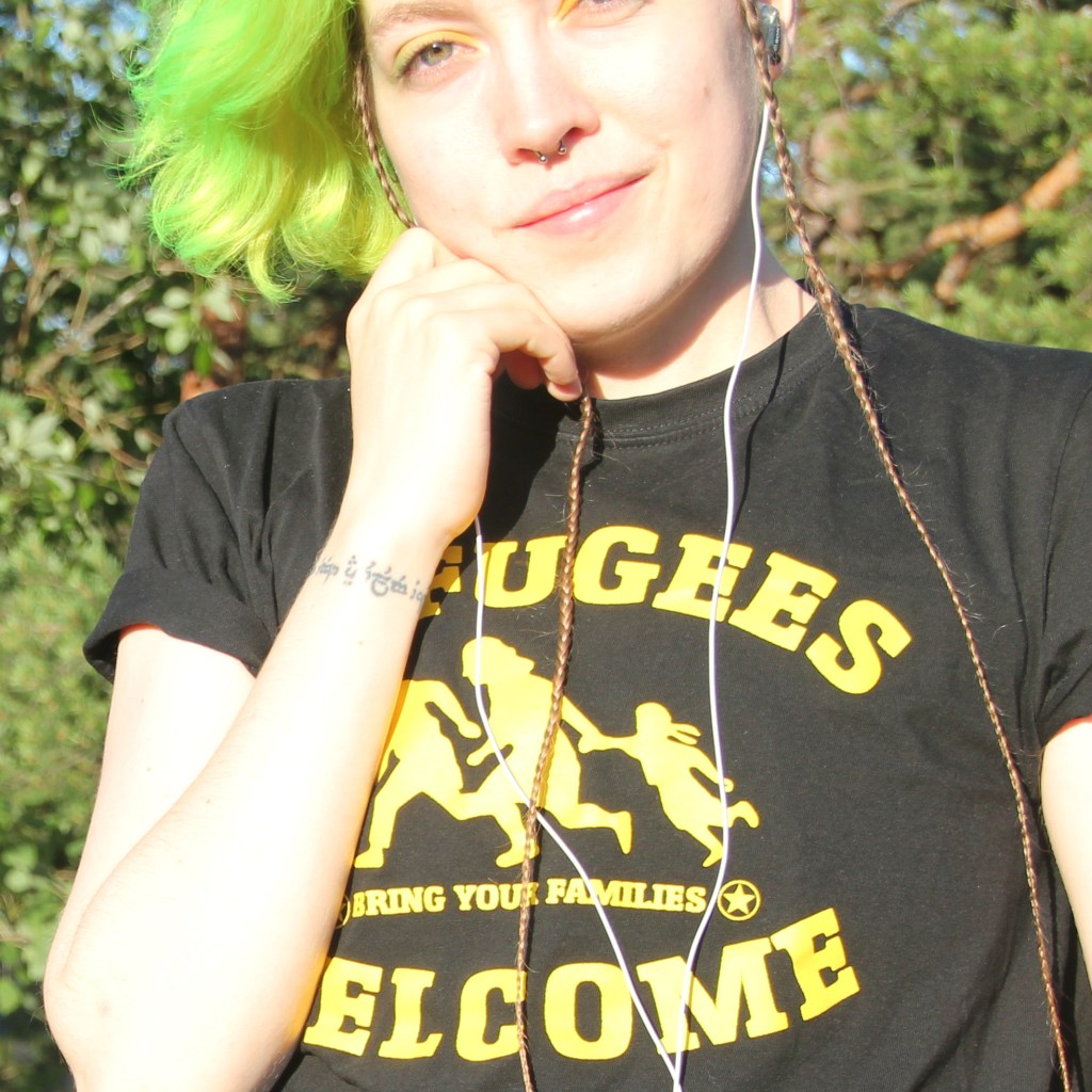 Photo of me: a white person with yellow green hair and eye shadow, and a black shirt with "Refugees welcome" written in yellow