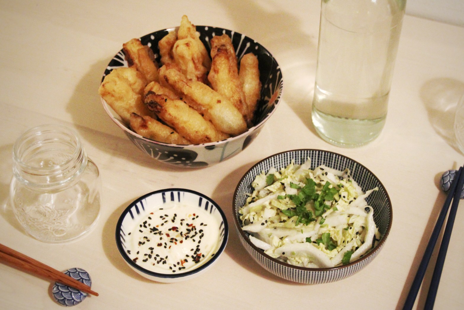 Three bowls containing, from left top to right bottom, pineapple fritters, mayo with sesame seeds, and coleslow. There's a glass and a water bottle next to them, as well as two pairs or chopsticks.